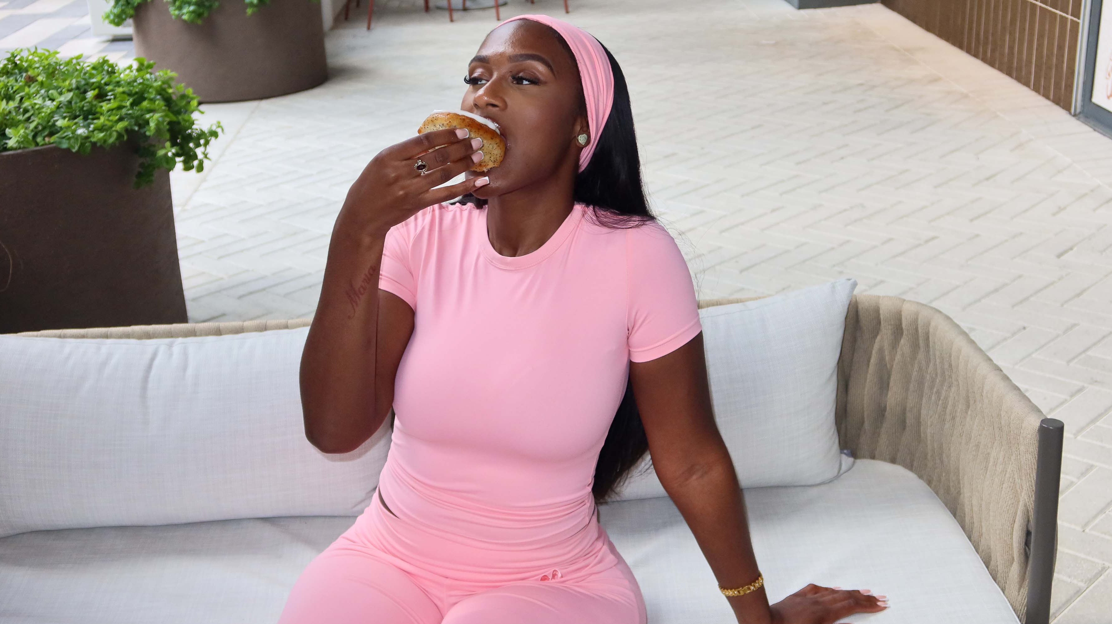 A woman in pink outfit sitting on a sofa eating a lemon cake outdoors.